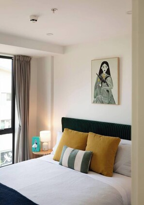 A hotel room with a bed, a nightstand, a lamp, a window with curtains, and a wall art piece. - Luxe 1BR City Apt | Modern, Spacious w/ Parking (Wellington)