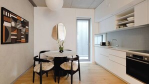 A modern kitchen with white cabinetry, a black oven, and a white countertop. There is a dining table with a mirror above it and a vase with flowers on the table. - Executive 2BR Designer Townhouse + Rooftop Oasis (Wellington)