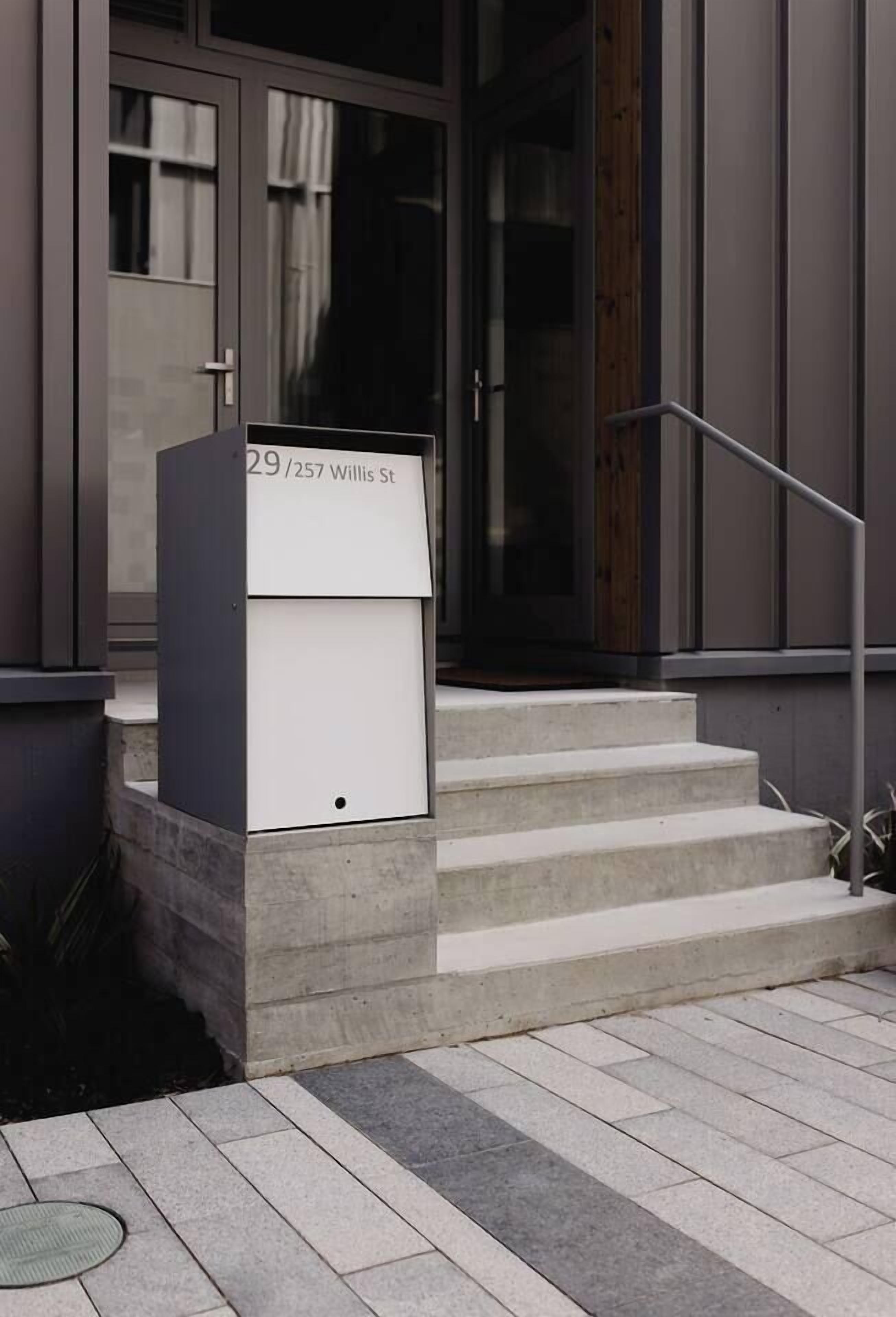 Une boîte aux lettres avec l’adresse « 29/257, rue Willis » est posée sur une marche en béton à l’extérieur d’un immeuble doté de grandes fenêtres et d’une rambarde métallique.