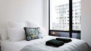 A hotel room with a bed, white bedding, a floral pillow, and a view of a building through a large window. - Chic 2BR Apartment | Secure Gated Complex in CBD (Wellington)