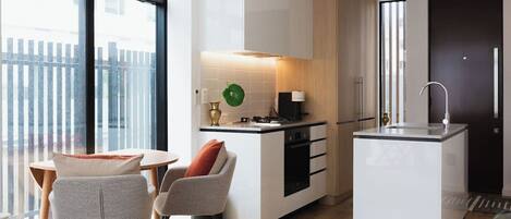 Et moderne køkken med en central ø, et spiseområde med to stole, og udsigt udendørs gennem store vinduer.