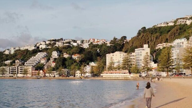 Une plage où une personne se promène le long du rivage, avec des bâtiments en arrière-plan et un ciel dégagé.