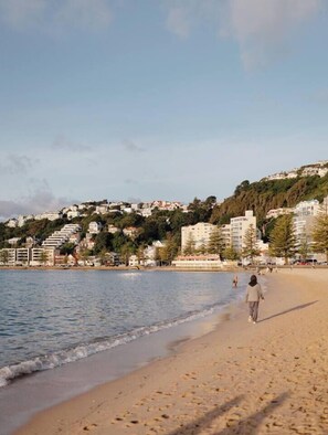 Une plage où une personne se promène le long du rivage, avec des bâtiments en arrière-plan et un ciel dégagé.