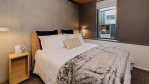 A bedroom with a bed, bedside table, lamp, and a window with a view of buildings. - Spacious 2BR Oasis in Central Wellington w Terrace (Auckland)