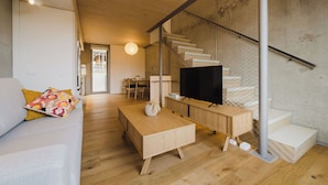 Un salon moderne avec un plancher en bois, un canapé agrémenté de coussins colorés, une table basse en bois, un téléviseur et un escalier doté d’une rampe en verre.