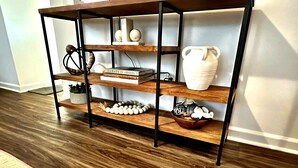 A wooden console table with black metal legs, featuring three shelves. The top shelf holds a white vase and a book titled 'Curate'. The middle shelf has a bowl with decorative items, and the bottom shelf displays a string of white beads. - Southern Solace (Newnan)