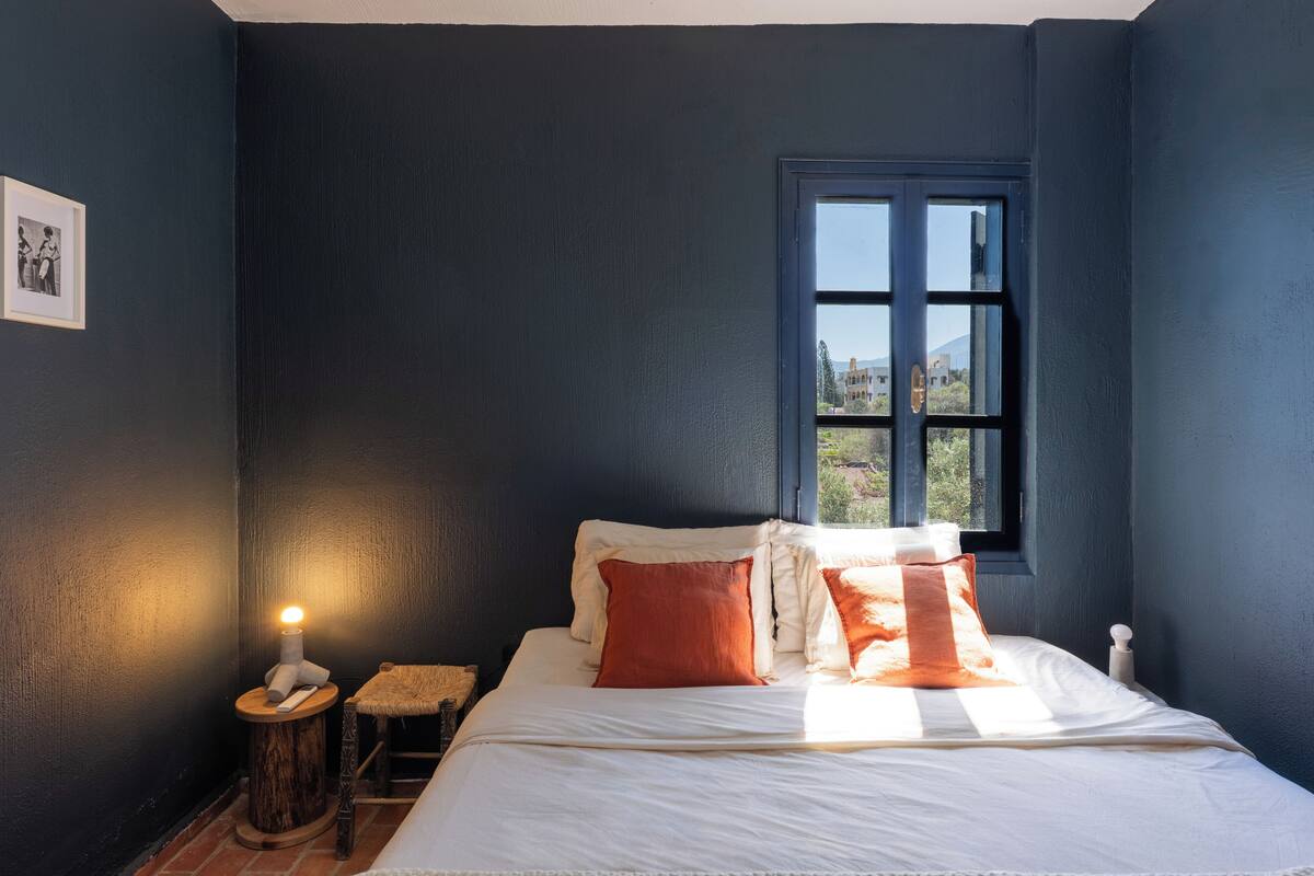 A bedroom with a bed, pillows, a nightstand, a lamp, a window, and a framed picture on the wall.