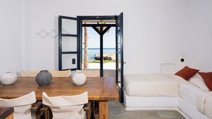 Une pièce avec une table à manger en bois, des chaises blanches, un lit avec une literie blanche et une vue sur la mer par une porte ouverte.