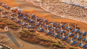 Vue aérienne d’une plage avec des rangées de transats et de parasols, une plage de sable et des vagues en arrière-plan.