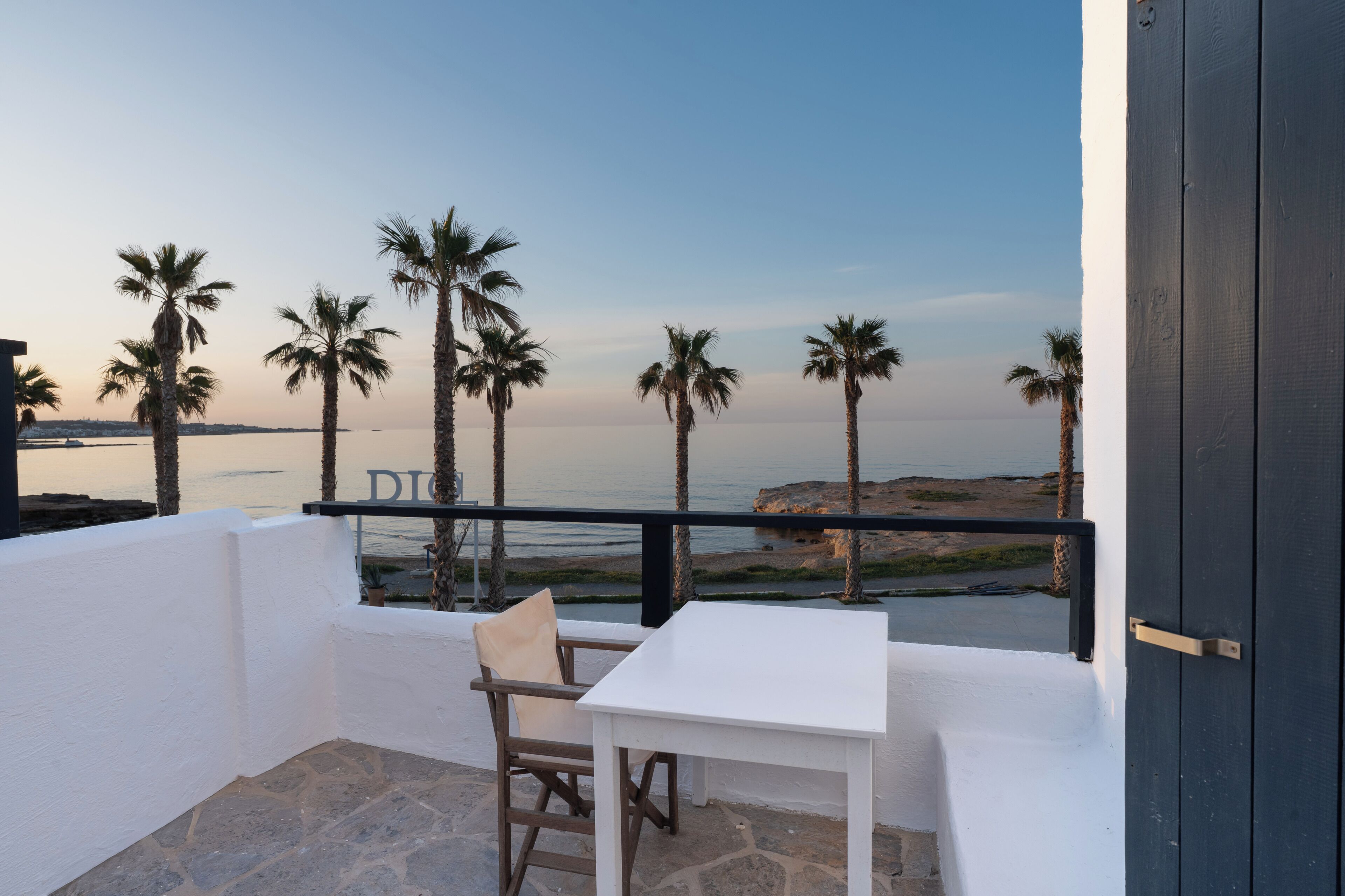 A balcony with a white table and chair, overlooking a beach with palm trees and a clear sky.