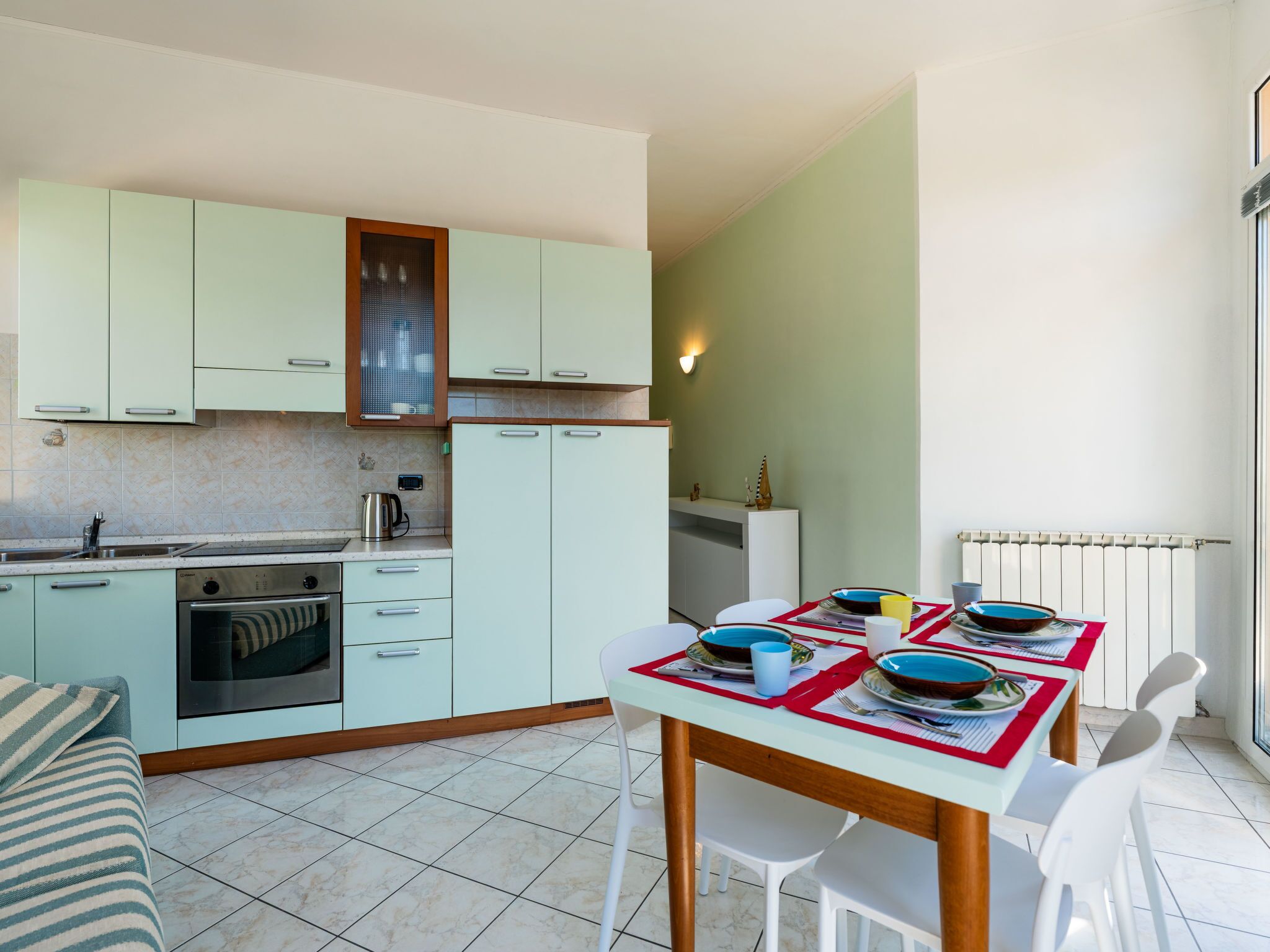 Een kleine keuken met lichtgroene kastjes, een inbouwoven en een eethoek met een tafel voor vier personen.