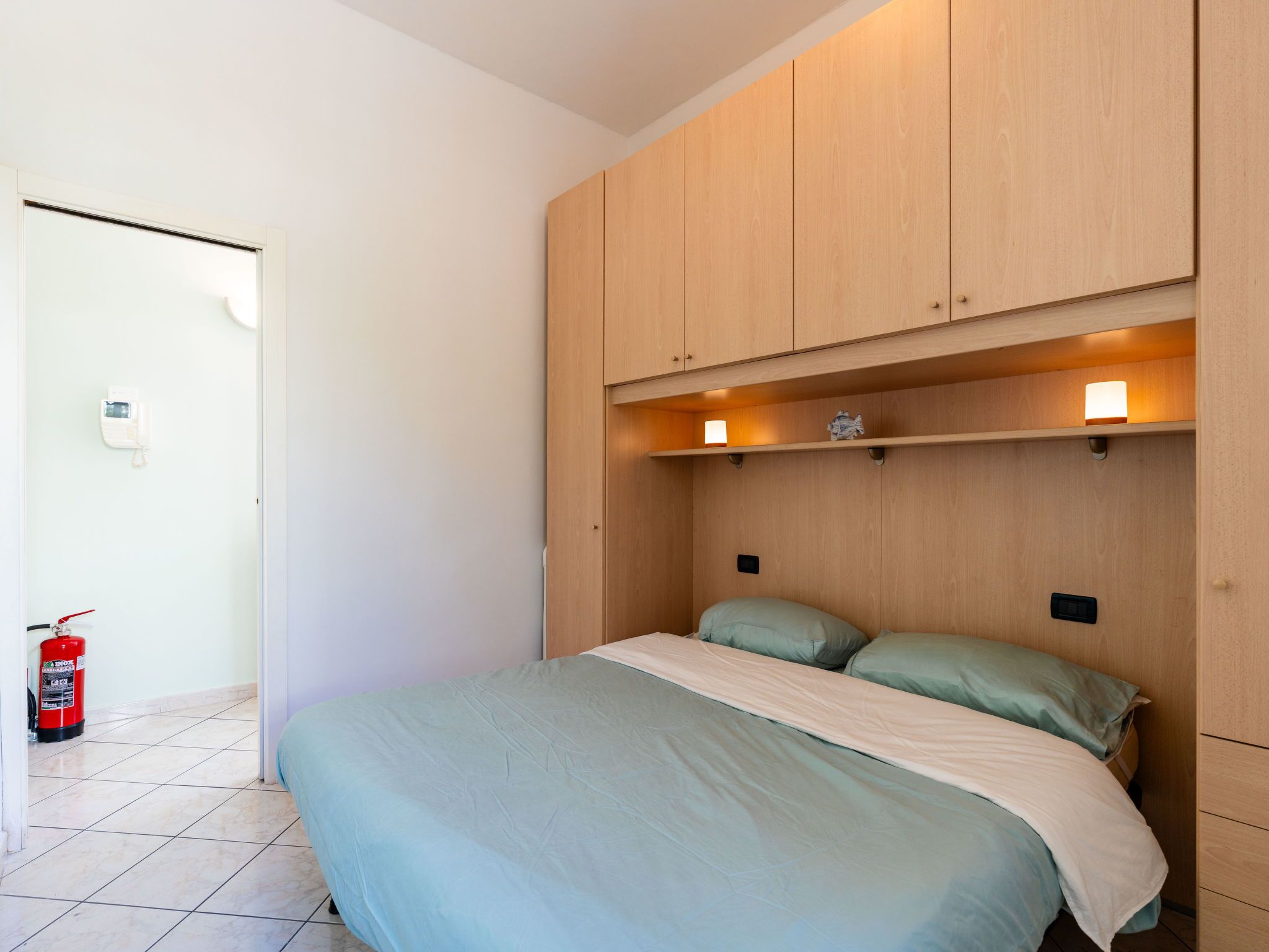 Een slaapkamer met een bed, een houten garderobe, bedlampen en een brandblusser.