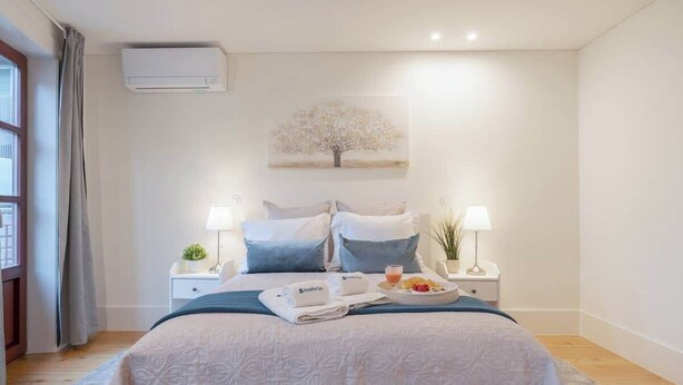 Un dormitorio ordenado con una cama, dos mesitas de noche, un aire acondicionado de pared, un cuadro de un árbol y una bandeja de desayuno sobre la cama.