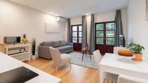 Una sala moderna con un sofá gris, una mesita redonda pequeña y una mesa de centro blanca. Hay un escritorio blanco con una computadora y una planta en maceta.