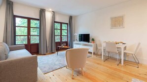 Una sala de estar moderna con un sofá, una mesa de comedor con sillas, un televisor y ventanas amplias con cortinas.