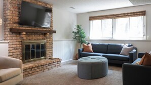 Ein Wohnzimmer mit Kamin aus Backsteinen, einem flachbildfernseher darüber sowie einem blauen Sofa und einem grauen Hocker.