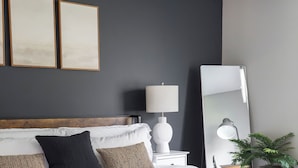 Ein ordentlich bezogenes Bett mit weißen Bettwäsche und einem hölzernen Kopfteil, ein Nachttisch mit Lampe, ein großer Spiegel und gerahmte Kunstwerke an der Wand.