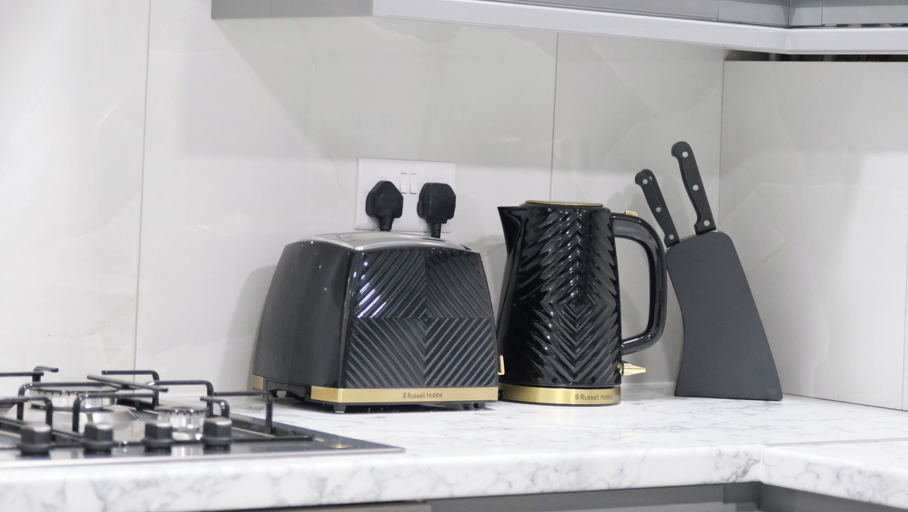 Une cuisine équipée d’une cuisinière à gaz, d’un grille-pain, d’une machine à café et d’un ensemble de couteaux posés sur un plan de travail en marbre.