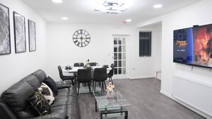 Un salon moderne avec une table basse en verre, un canapé en cuir, un coin repas avec une table blanche et des chaises noires, une horloge murale et un téléviseur diffusant le film « Indiana Jones ».