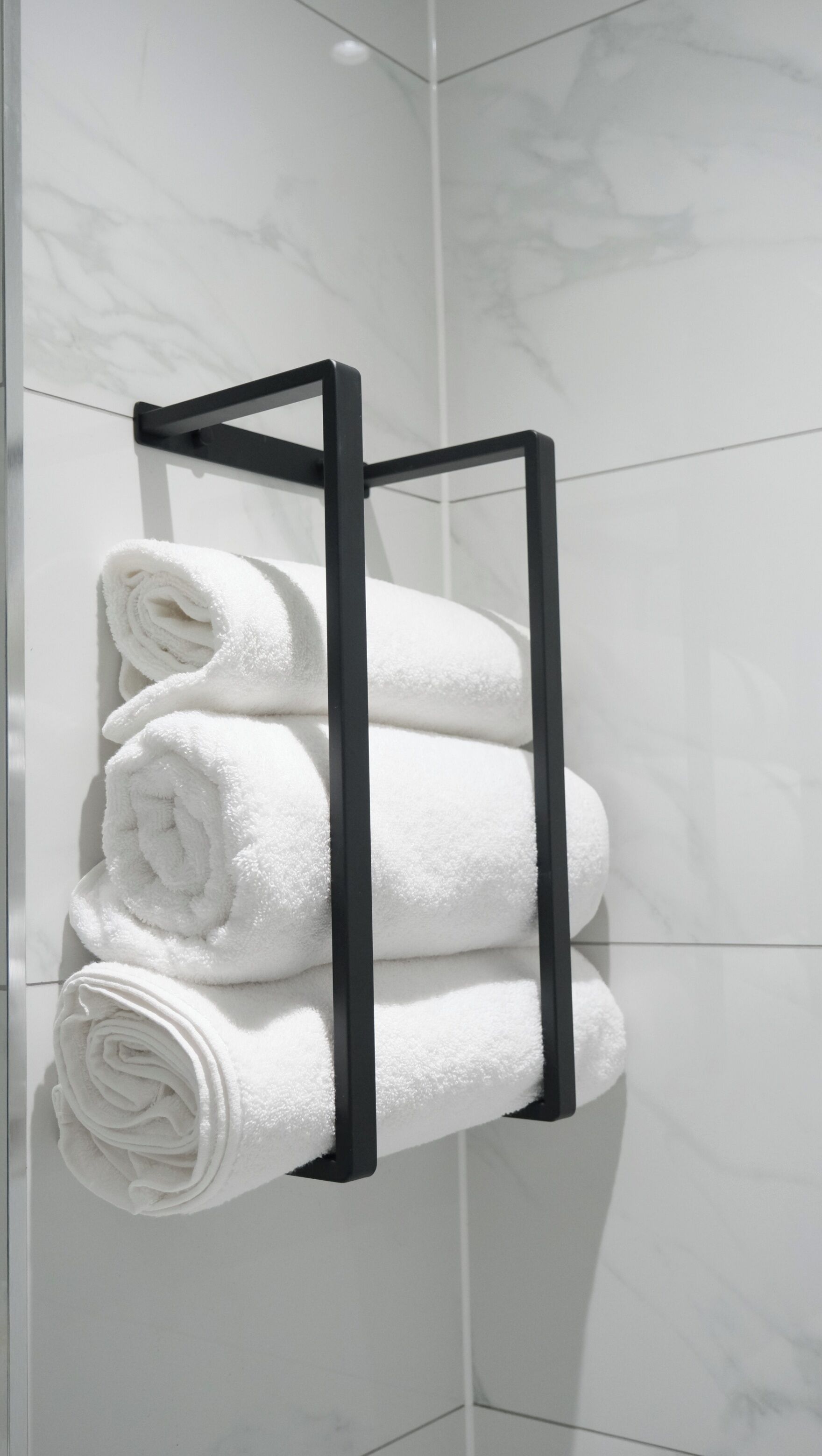 A black towel rack holding four neatly rolled white towels against a white tiled wall.