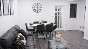 Un salon moderne avec un canapé en cuir noir, une table basse en verre et un coin repas comprenant une table blanche et des chaises noires. Une grande horloge murale est visible au-dessus de la table à manger.