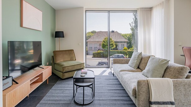 A modern living room with a flat-screen TV, a sofa, a small table, and a chair. There is a large window with a view of a house and trees. - Blueground | Senningerberg, balcony, nr Parc de Merl (Luxembourg)