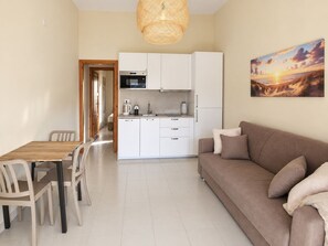 A modern kitchen with white cabinets, a dining table with chairs, a sofa with cushions, and a painting of a sunset on the wall.