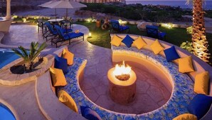 Un espace extérieur circulaire aménagé avec des sièges, doté d’un foyer, entouré de canapés et de fauteuils, et offrant une vue sur une piscine et un jardin paysager.