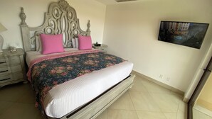 Une chambre à coucher avec un grand lit, une tête de lit décorative, des coussins roses, une couvre-lit à motifs floraux, une table de chevet avec une lampe et un téléviseur fixé au mur.
