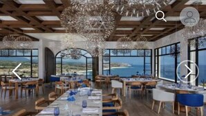 Une salle à manger spacieuse avec de longues tables dressées pour le repas, de grandes fenêtres offrant une vue sur la mer et un lustre moderne.