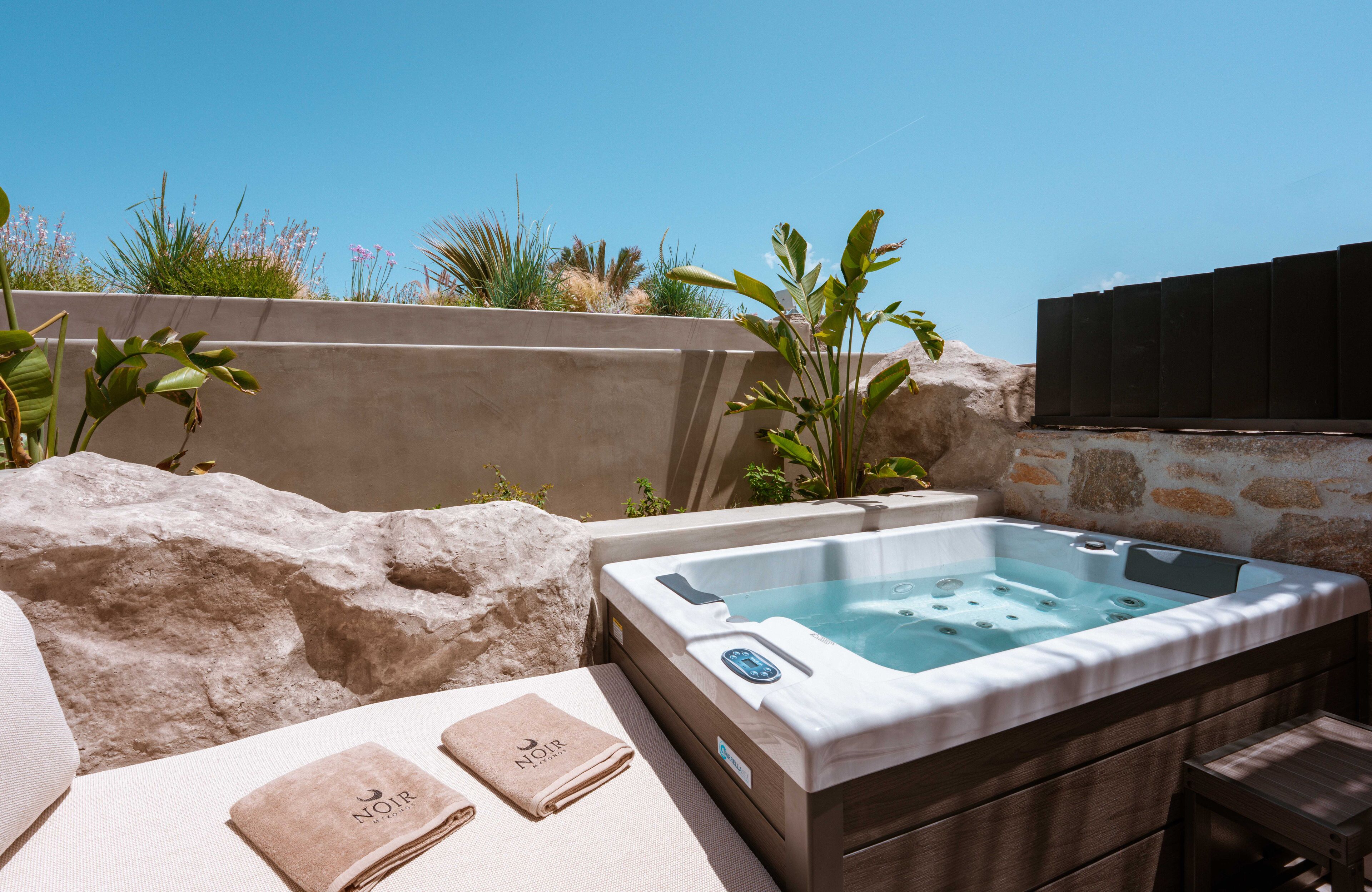 A hot tub with a stone wall backdrop, a lounge area with towels, and a clear blue sky.
