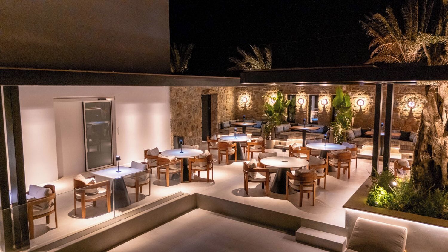 An outdoor dining area with multiple round tables and chairs, surrounded by stone walls and palm trees.