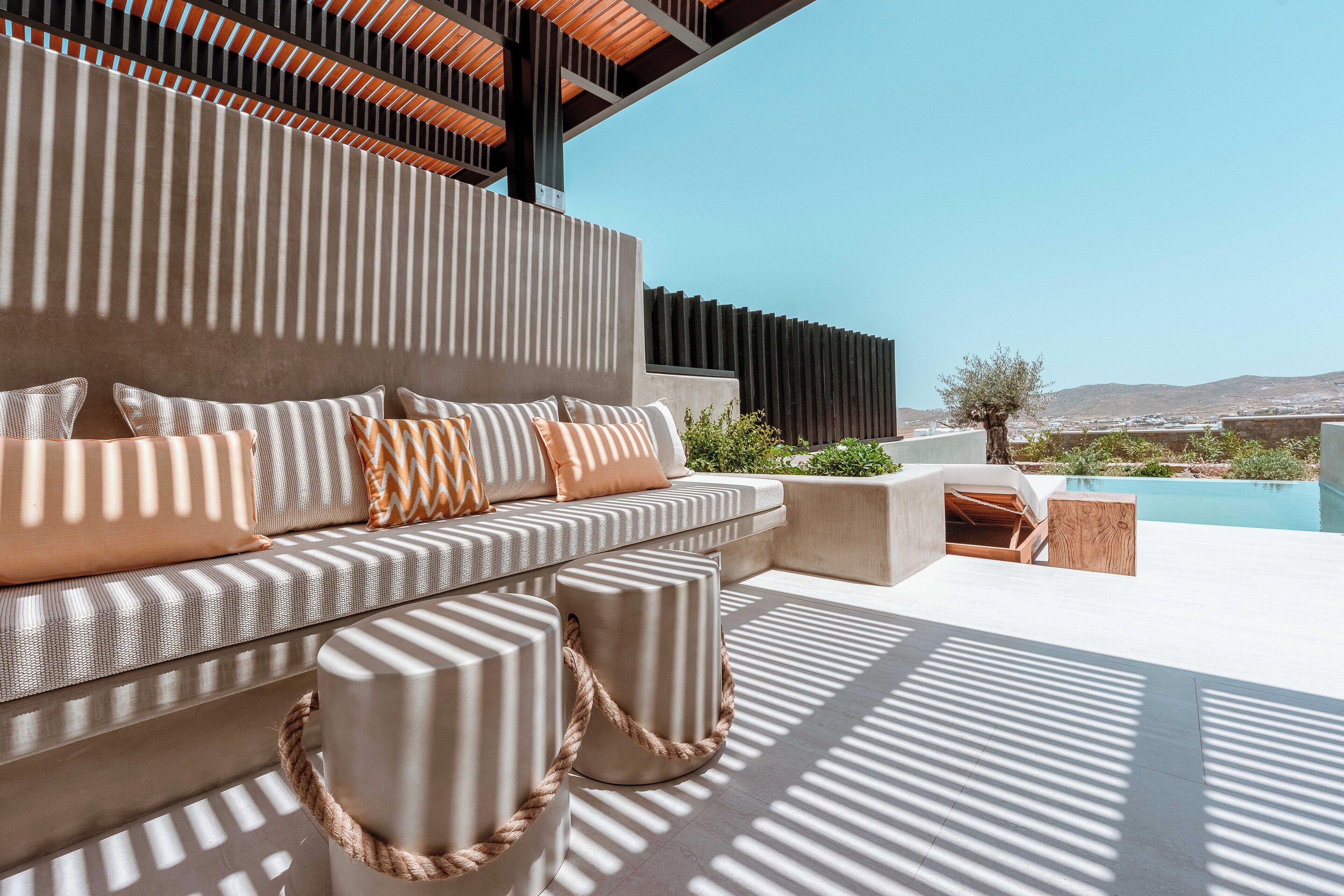 A modern outdoor seating area with a sofa, ottomans, and a wooden side table, overlooking a pool and a scenic landscape.