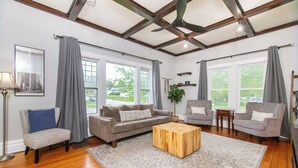 A spacious living room with hardwood floors, a large gray patterned rug, and a wooden coffee table. There are three sofas and a chair, with a large window providing a view of the outdoors. - Charming 1950's Home Near Downtown, Fenced Yard, Pet Friendly! (Rapid City)