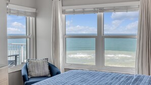 Un dormitorio con una cama grande, una silla y vistas al océano a través de dos ventanas.