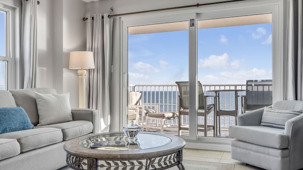 Una sala moderna con sofá, sillón y una mesa de centro de vidrio. Un amplio ventanal corredero da a un balcón con vistas al océano.