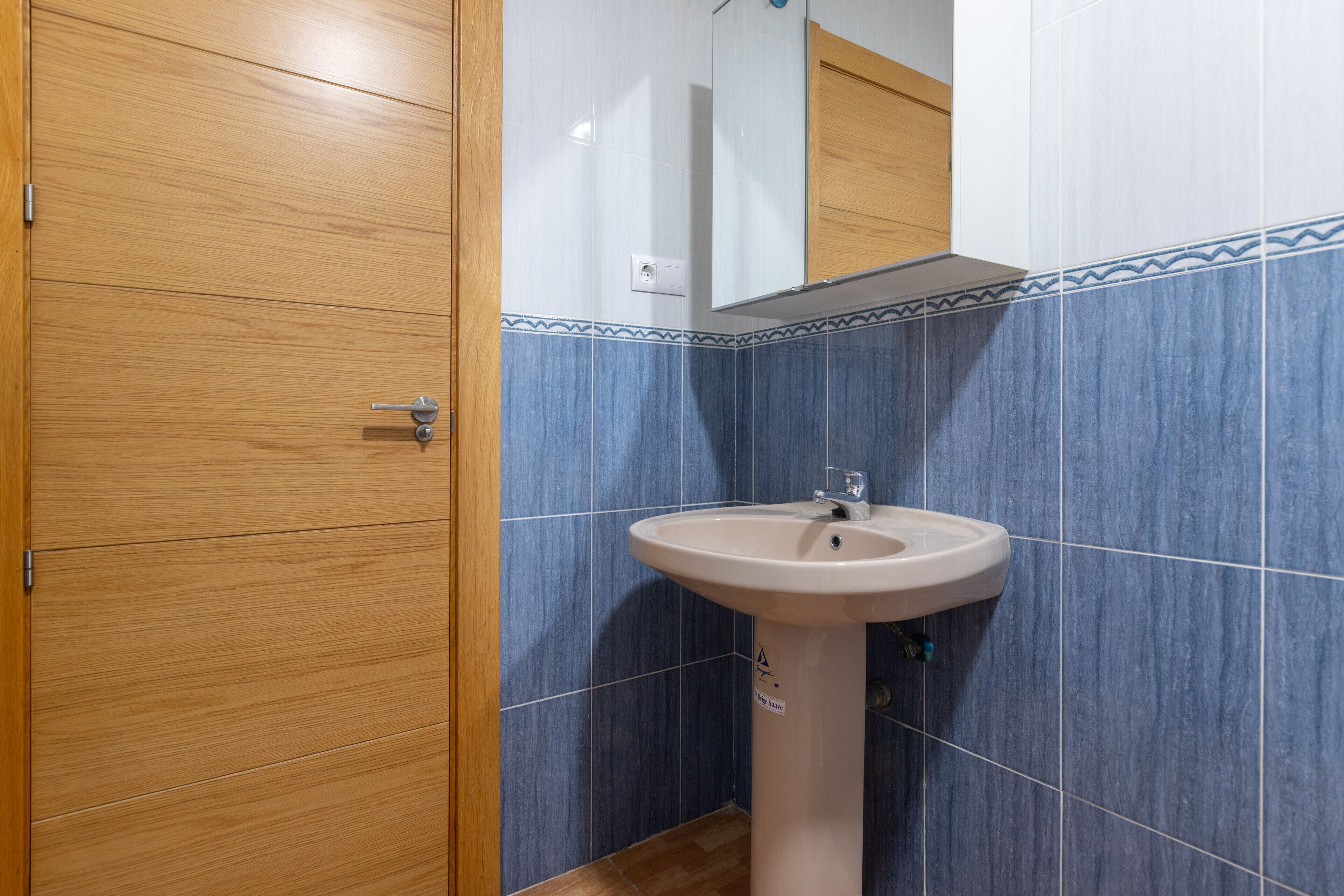 Une salle de bain avec une porte en bois, un lavabo surmonté d’un miroir et des murs recouverts de carreaux bleus.
