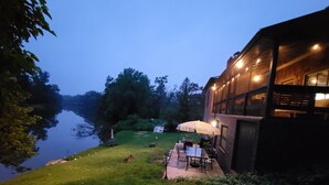 En terrasse ved floden med udendørs møbler og et bord under en parasol, ved siden af en bygning med store vinduer og lyskæder.