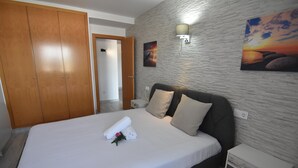 Un letto rifatto con cura, un asciugamano arrotolato e un fiore, un divano grigio con cuscini, un armadio in legno e una lampada fissata al muro.