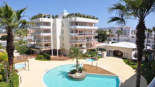 Un hotel su più piani con area piscina, palme e un cielo azzurro limpido.