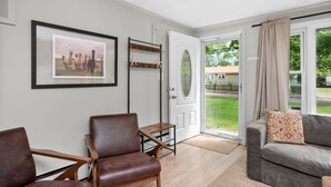 Un salon avec un fauteuil en cuir marron, un canapé gris, un tableau encadré au mur et une porte donnant sur un jardin.