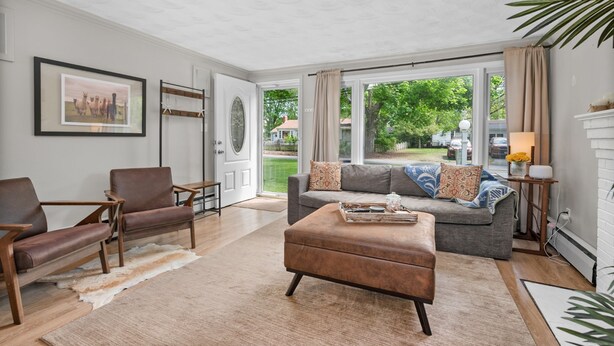 Un salon avec un canapé gris, un fauteuil en cuir marron et un pouf. On y trouve une cheminée, une lampe et un tableau encadré au mur.