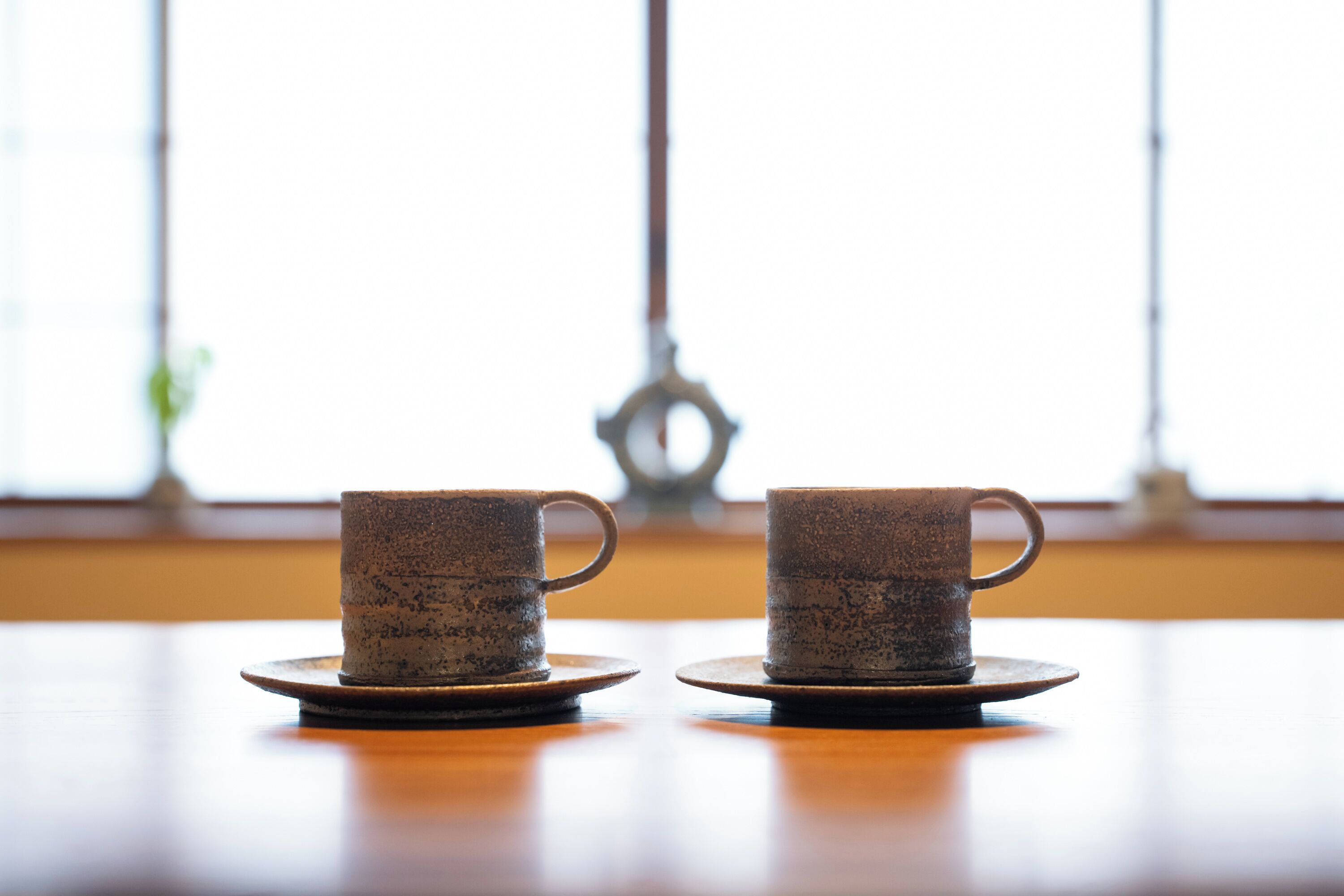 Two ceramic cups on saucers on a wooden table with a window in the background.