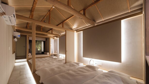 Une pièce avec un plafond haut, des poutres en bois, un ventilateur de plafond et deux lits avec du linge de lit blanc.