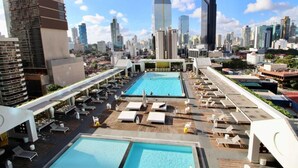 A rooftop pool area with multiple swimming pools, lounge chairs, and a city skyline in the background.
