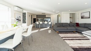 A modern living room with a grey sectional sofa, a dining area with white chairs, and a kitchen in the background.