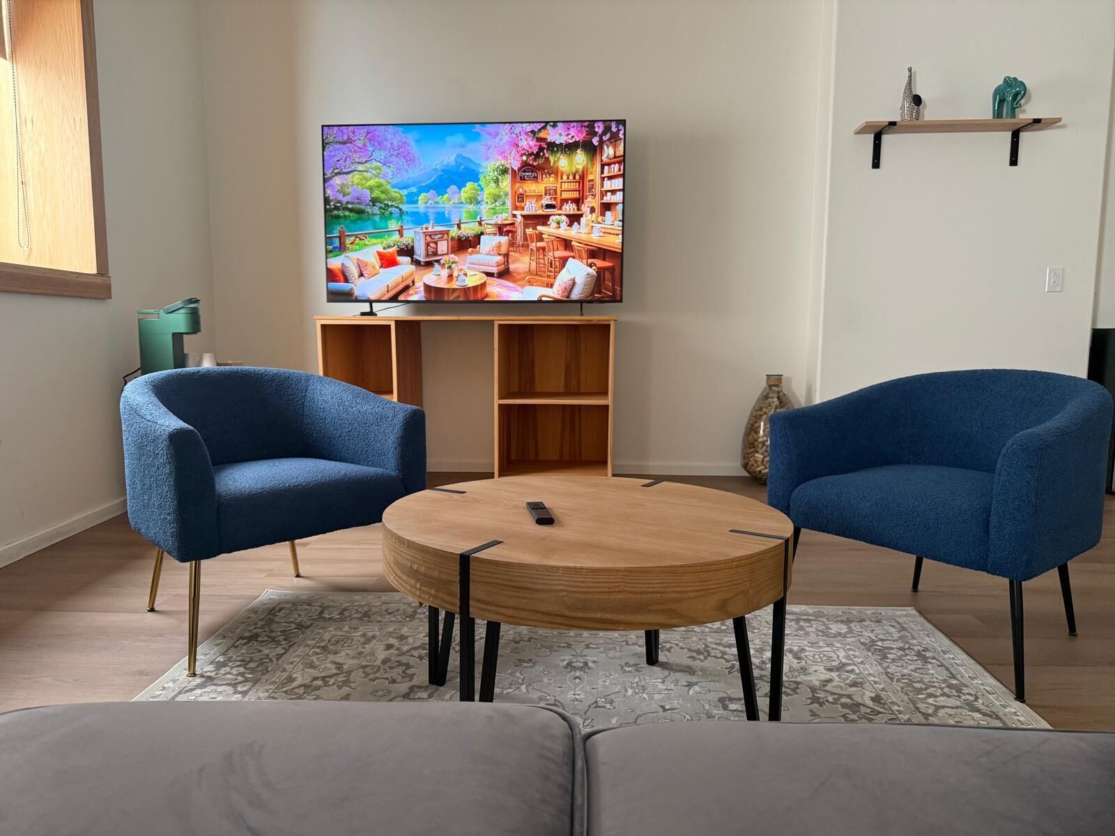 A living room with a TV, two blue armchairs, a round coffee table, and a bookshelf.