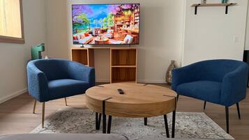 A living room with a TV, two blue armchairs, a round coffee table, and a bookshelf.