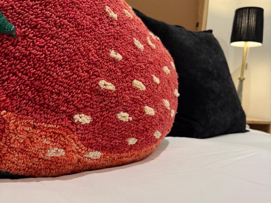 Un almohadón grande con forma de fresa, de textura marcada, sobre una cama con sábana blanca.