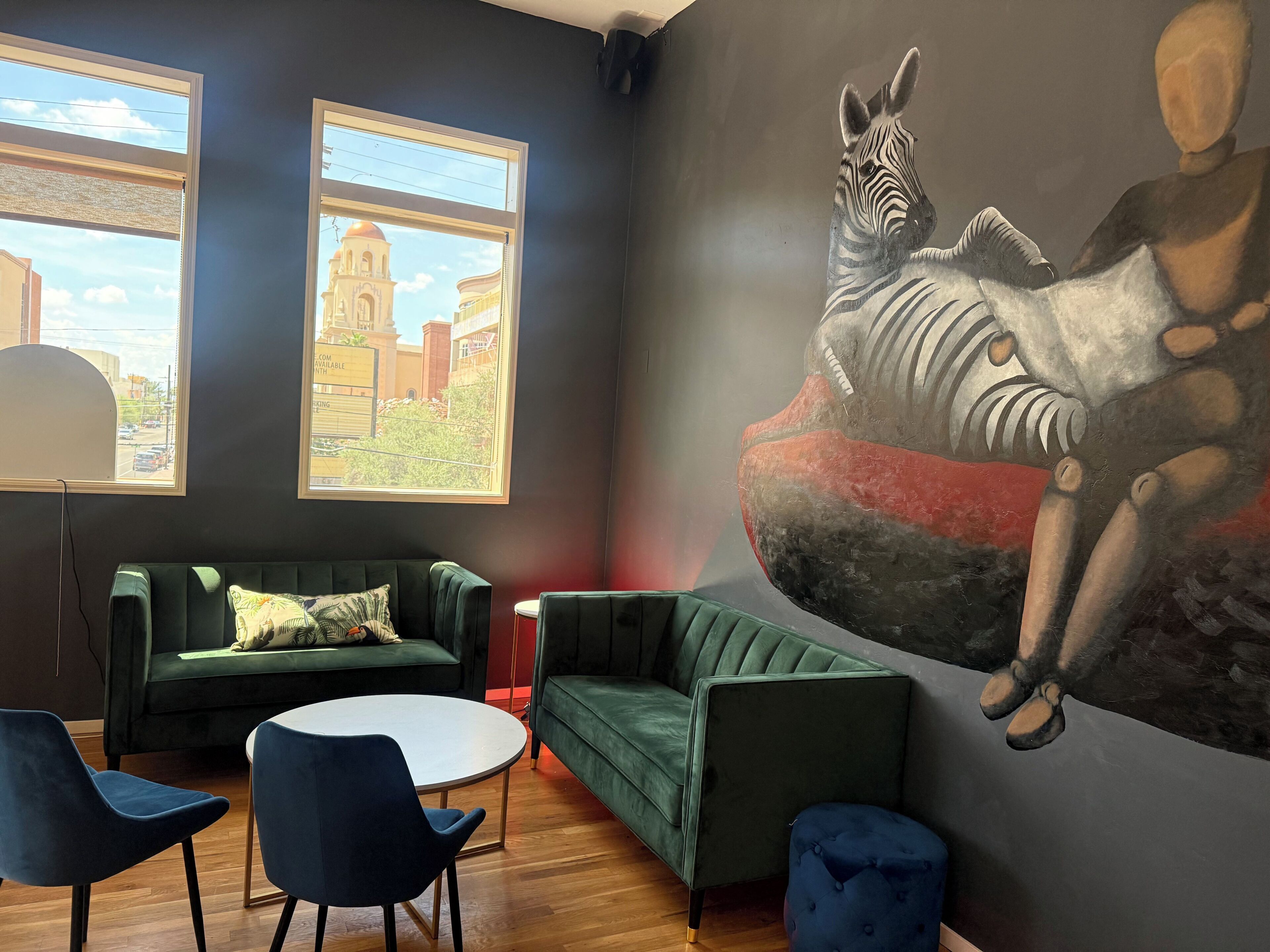 Ein Zimmer mit einer großen Zebradarstellung, zwei Fenstern mit Blick auf Gebäude, einem grünen Sofa, einem runden Tisch und zwei blauen Stühlen.